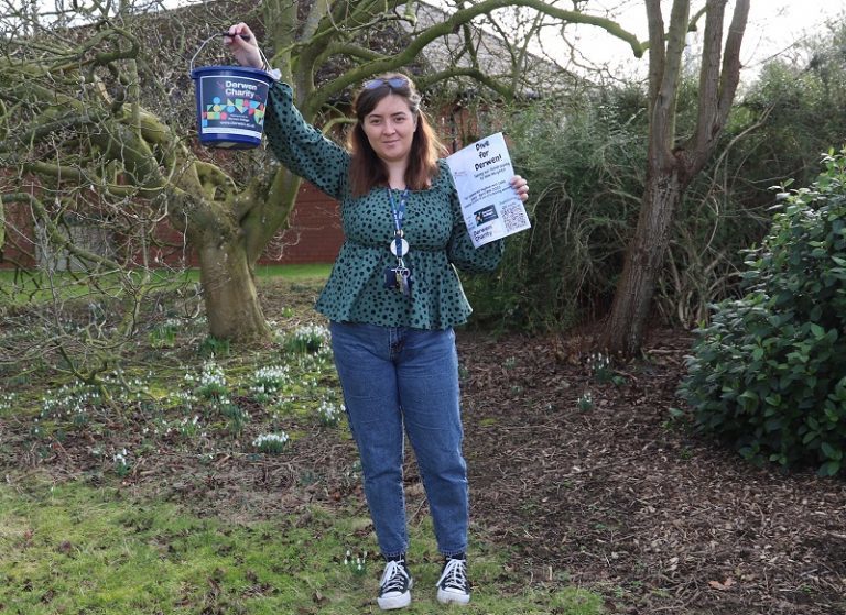 Derwen Learning Support Assistant Tia - pictured outside amongst the snowdrops - holds up a Derwen Charity collection bucket and promotional poster.