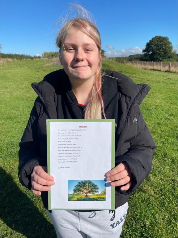 Student Tiff stood outside in the breezy countryside wearing a coat and holding up her winning poem about an oak tree.